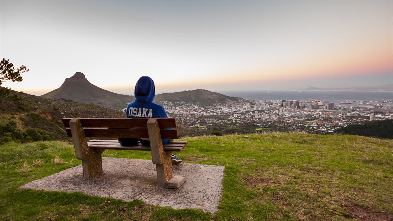 A time-lapse of a man sitting on a bench wearing a hoodie, watching the sunset over the City of Cape Town