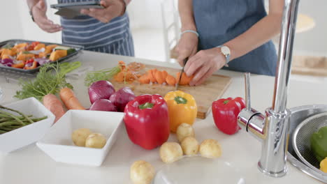 Diverse-couple-wearing-aprons-and-cooking-with-tablet-at-home,-slow-motion