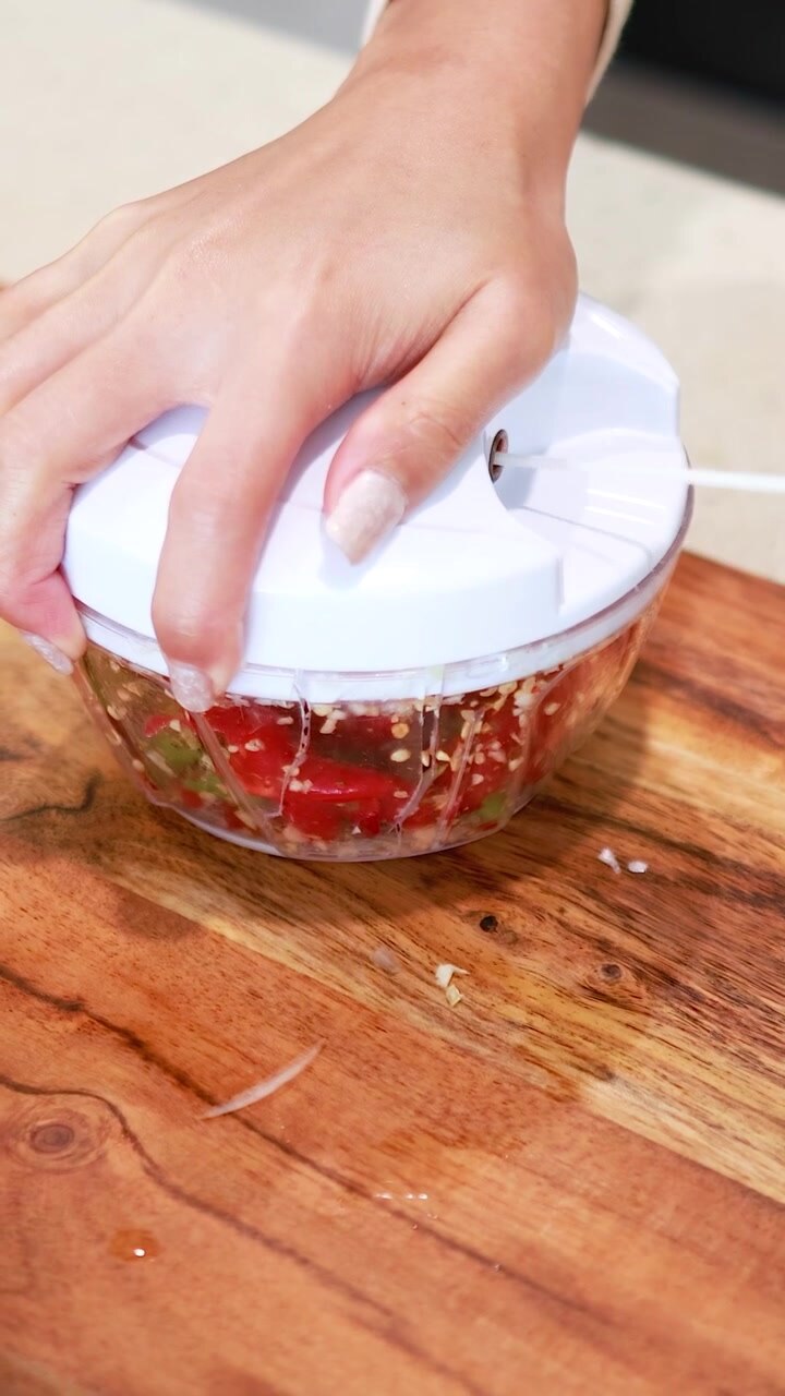 Una mano aziona un tritatutto manuale su un tagliere di legno, tritando efficientemente aglio e peperoncino in una cucina ben illuminata