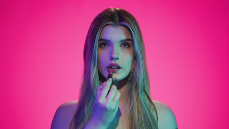 medium slow motion shot of a young attractive and beautiful woman putting red lipstick on her lips and giving a lewd look to the camera in front of pink background