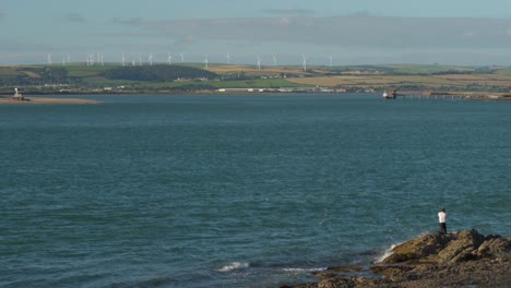 A-fisherman-and-a-wind-farm-occupy-two-sides-of-a-coastline-on-a-bright-summer-day