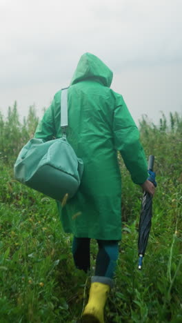 vista trasera de una mujer con un impermeable verde que lleva una bolsa de color menta y un paraguas, con botas de lluvia amarillas, caminando con precaución por un camino rodeado de vegetación alta y plantas
