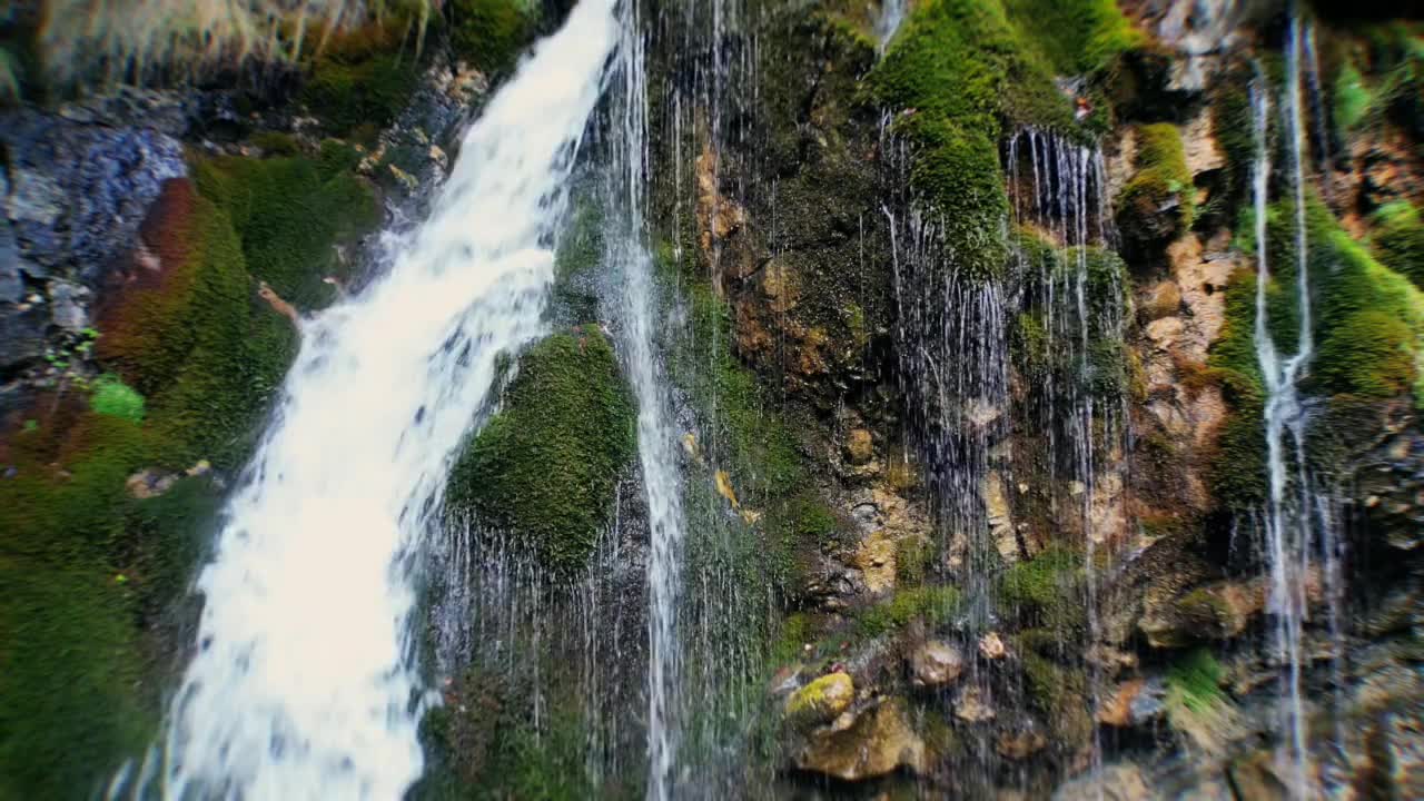Static close-up drone view at waterfall in Bucegi National Park, Romania, showing tiny water streams flowing from moss-covered rocks, with a larger waterfall nearby