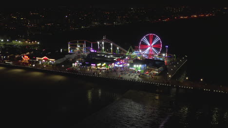 AERIAL:-Side-view-of-Santa-Monica-Pier-at-night-with-Ferris-Wheel-and-colorful-lights,