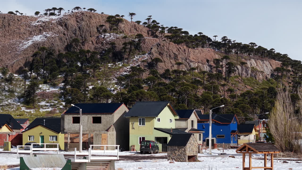 Aerial pan of Caviahue, Argentina, showing colorful houses in snow, set against rocky Andean hills and Araucaria forests under clear mountain skies