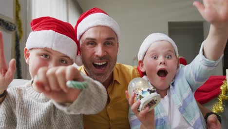 Portrait-of-caucasian-father-and-two-sons-wearing-santa-hats-waving-and-smiling-sitting-on-the-couch