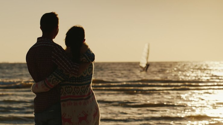 Jeune couple amoureux s'étreignant, regardant la mer et le coucher de soleil. Voilier flottant sur la mer, vue de dos.