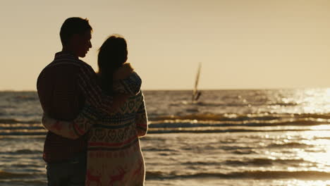 Young-Couple-In-Love-Hugging-Looking-At-The-Sea-And-The-Sunset-Sea-Sailboat-Floats-Back-View