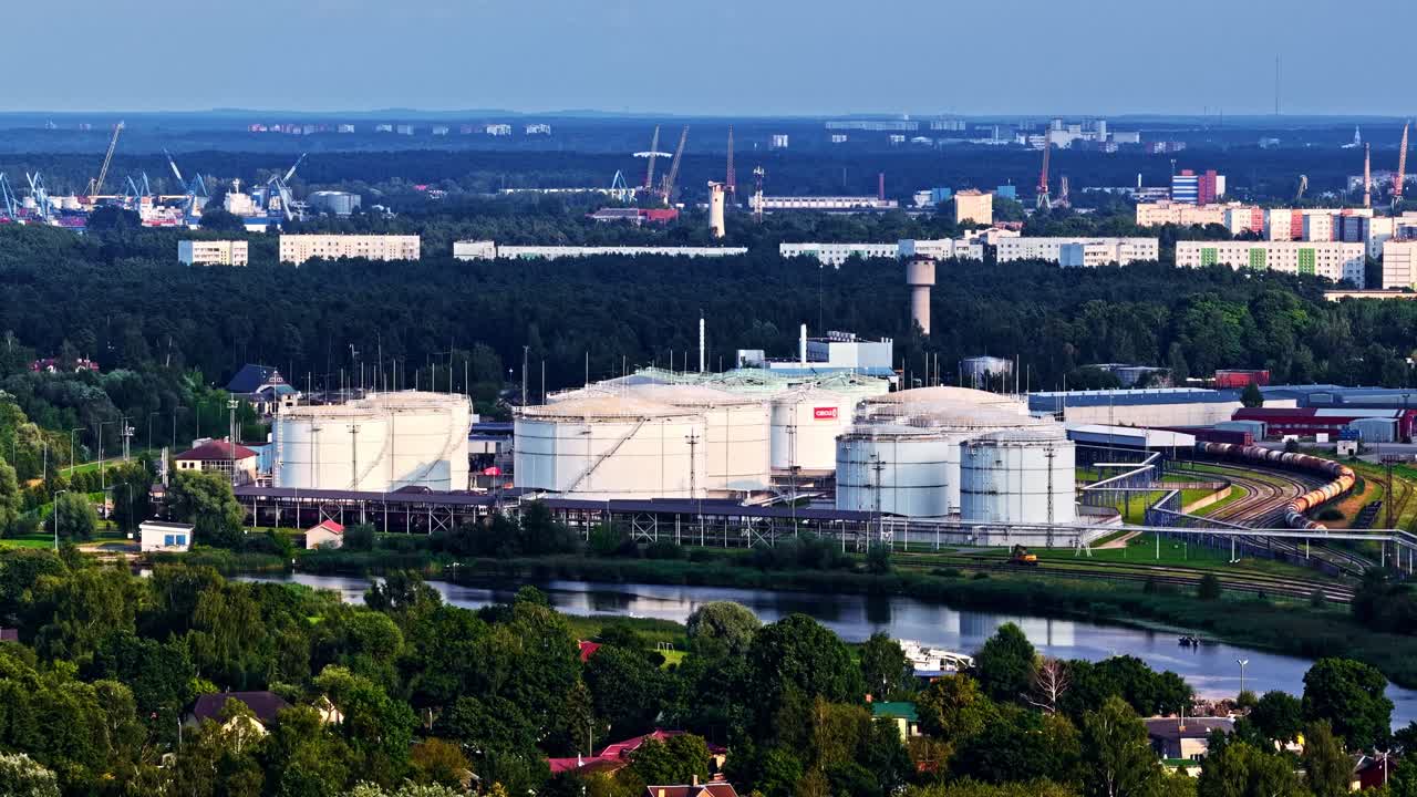 A wide aerial shot shows the large, white storage tanks of the Neste Latvija oil and fuel terminal, a key energy logistics hub located in the industrial Port of Riga, Latvia