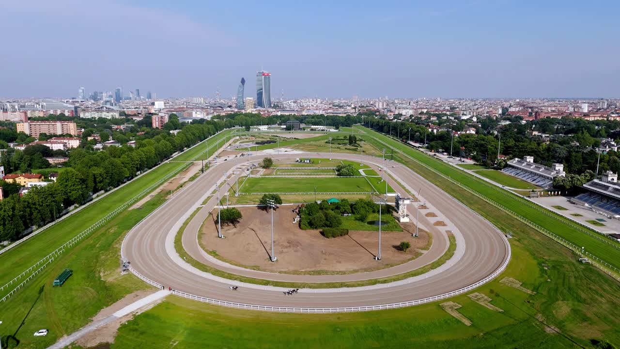 Wide-angle aerial view of horses trotting on a Milan racecourse, captured by drone, showing motion, city skyline, and expansive racetrack from above