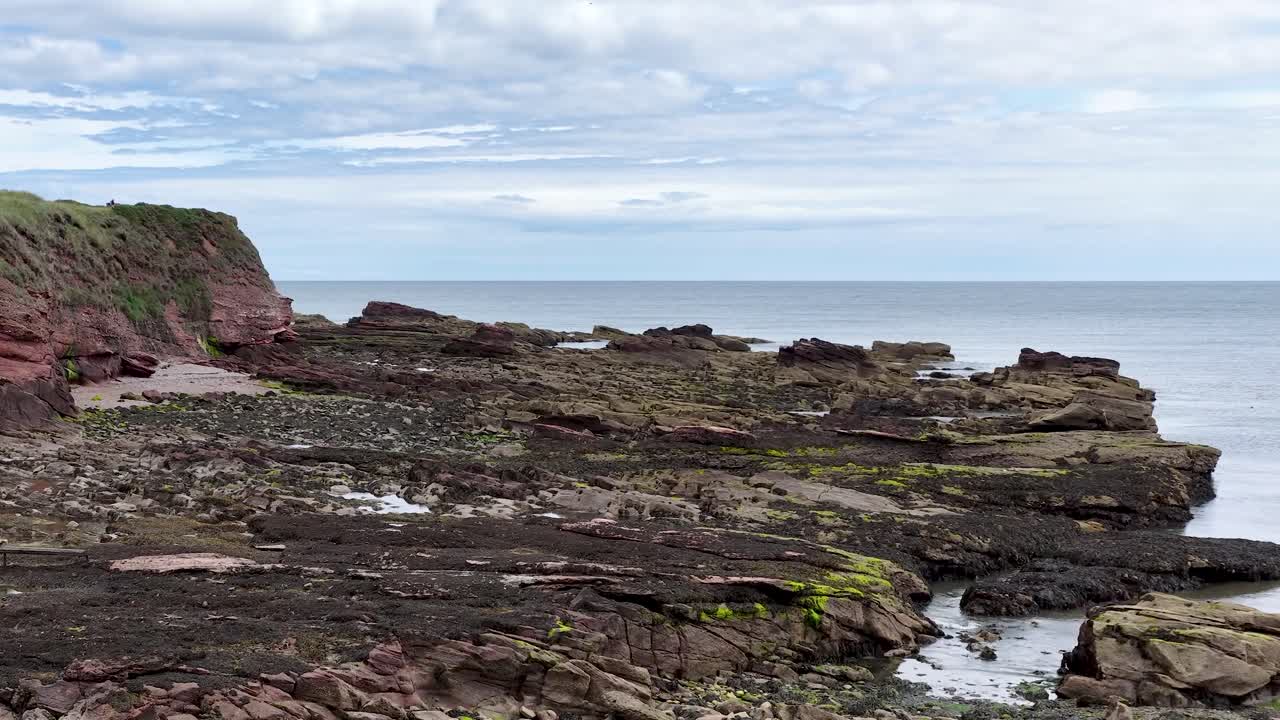 Camera slowly pans over red sandstone cliffs, rocky shore, and seaweed at low tide