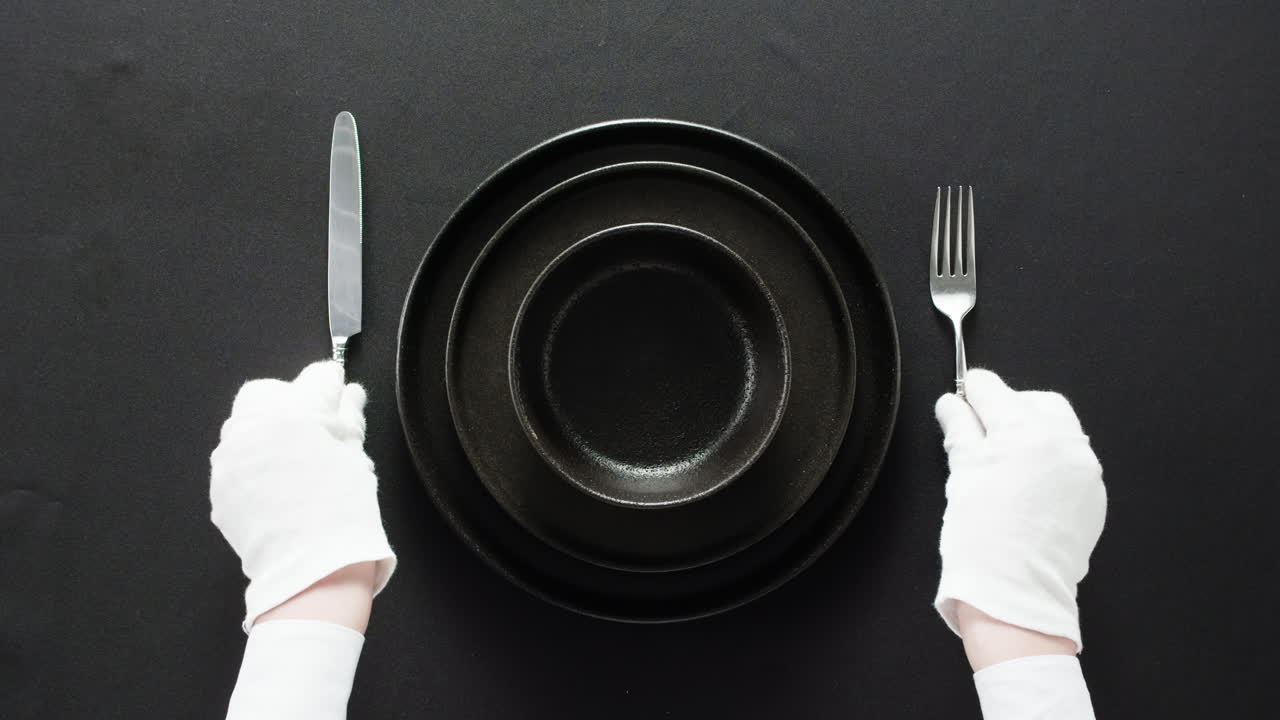Minimalist table setting with white plate, fork, and knife arranged neatly, and a gloved hand adjusting cutlery.