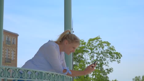 A-blonde-woman-with-her-hair-tied-back-under-a-metal-kiosk-uses-her-smartphone-and-smiles