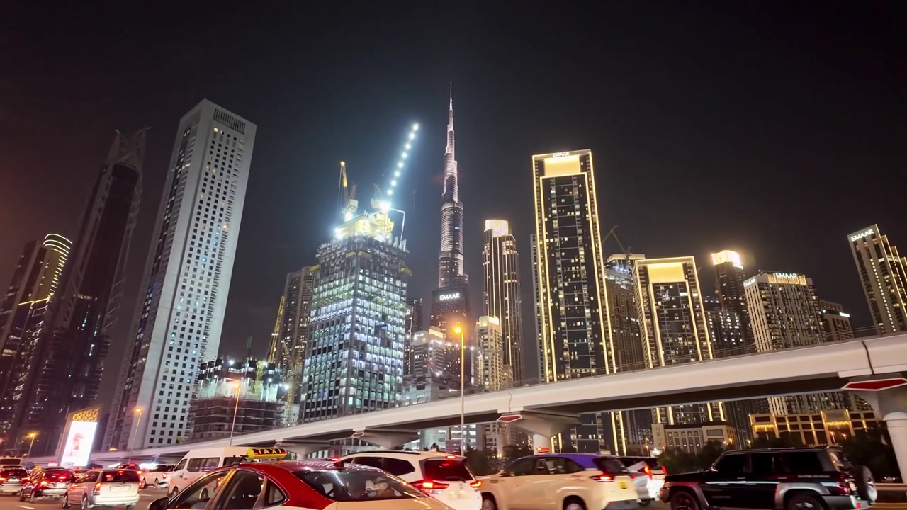 Busy Dubai night scene featuring illuminated skyscrapers, highway traffic, elevated roadway, and modern urban skyline with Burj Khalifa in background