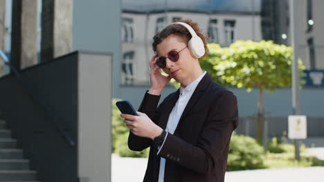happy joyful businessman listens to music on wireless headphones while walking on street near office