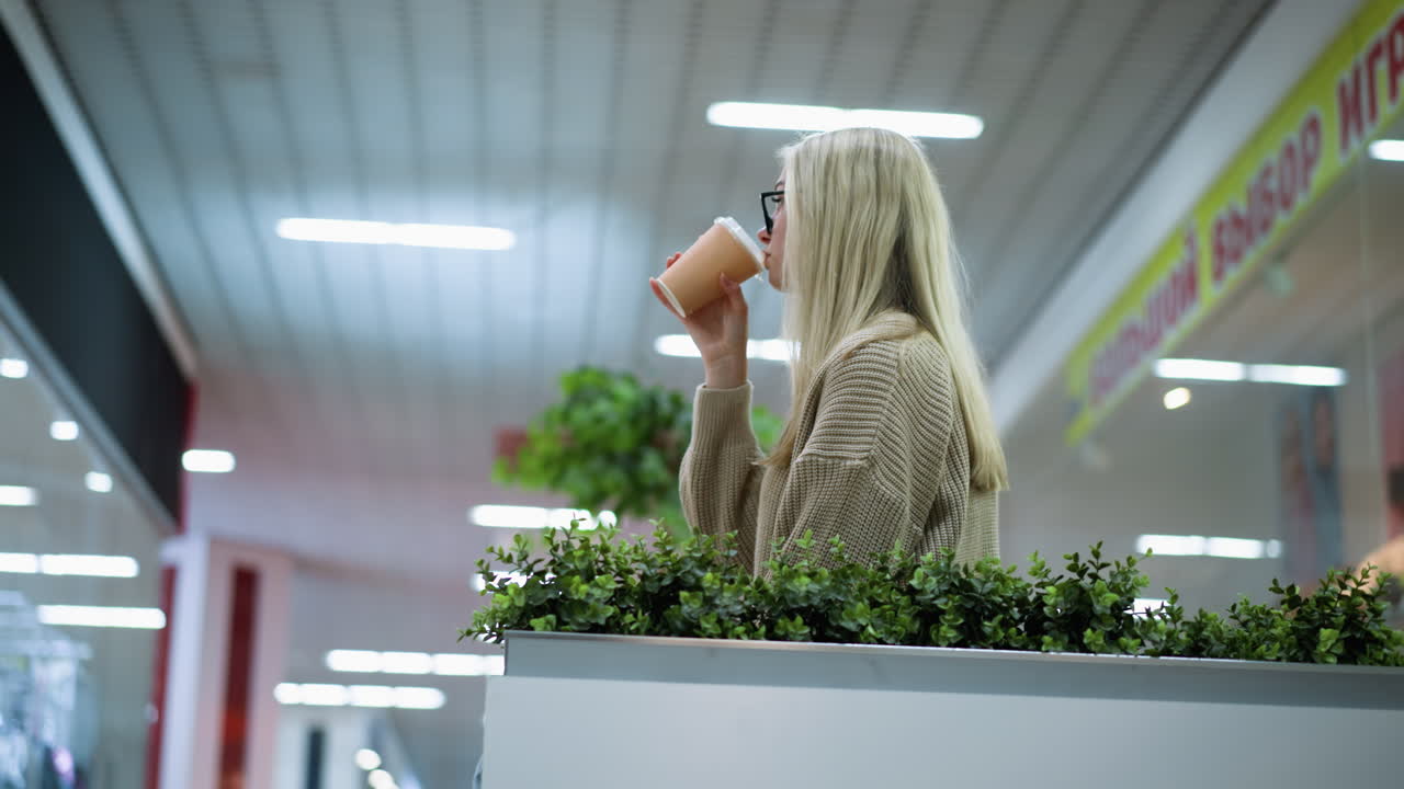 Mulher profissional saboreando café pensativamente com plantas decorativas por perto, o fundo desfocado da loja apresenta luzes decorativas brilhantes piscando