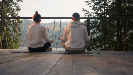 couple sits in lotus poses on terrace in early morning. concentrated young people do yoga exercise relaxing together in glamping on lake bank. healthy weekend