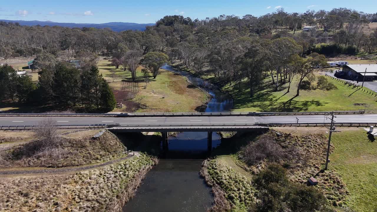 Drone footage moves steadily over a country bridge spanning a winding stream, surrounded by grassy fields, trees, and rural landscape in bright daylight