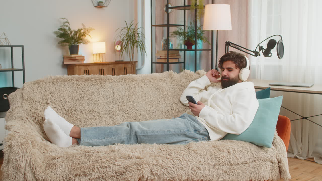 joven feliz y encantado con auriculares inalámbricos usando un teléfono inteligente relajándose sentado en el sofá en casa