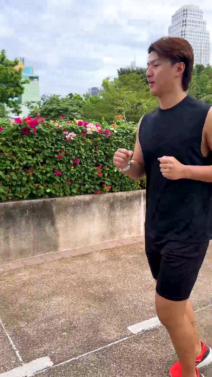 Pria bugar jogging di taman kota pada pagi berawan, kamera melacak dari samping, dengan pencahayaan alami.