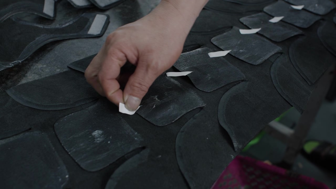 Worker placing adhesive strips on black shoe material pieces in factory setting