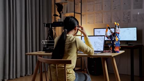 back view of teen asian girl with 3d printing having a headache while designing a cyborg hand on a laptop at home