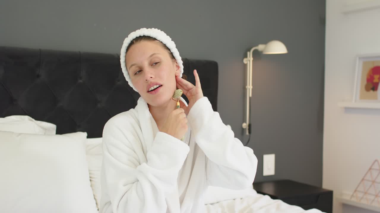 Erwachsene schwarze Frau im weißen Bademantel hält Gesichtsroller und massiert Kiefer bis Wange zur Hautpflege im Bett.