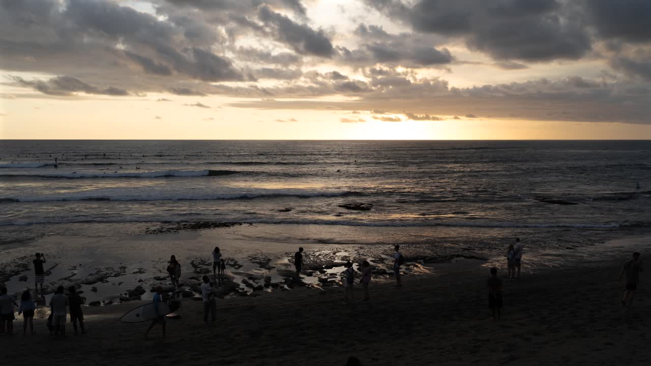 Indonesia, Bali, people in motion by the beach during sunset and tide, clouds move towards to the land and colors of the sky and clouds change from white to yellow to orange and pink finally black.