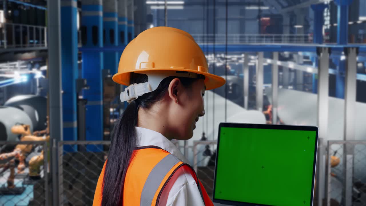 안전 헬을 입은 아시아 여성 엔지니어가 풍력 터빈 제조 공장에서 서서 녹색 화면 노트북을 보고 로 팔이 작동하는 동안 주위를 둘러보고 있습니다.