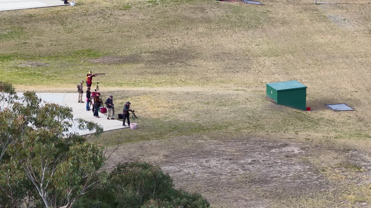 A group of people participate in clay target shooting at an outdoor range on a sunny day, viewed from an elevated, wide-angle perspective