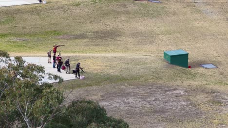 a group of people participate in clay target shooting at an outdoor range on a sunny day, viewed from an elevated, wide-angle perspective