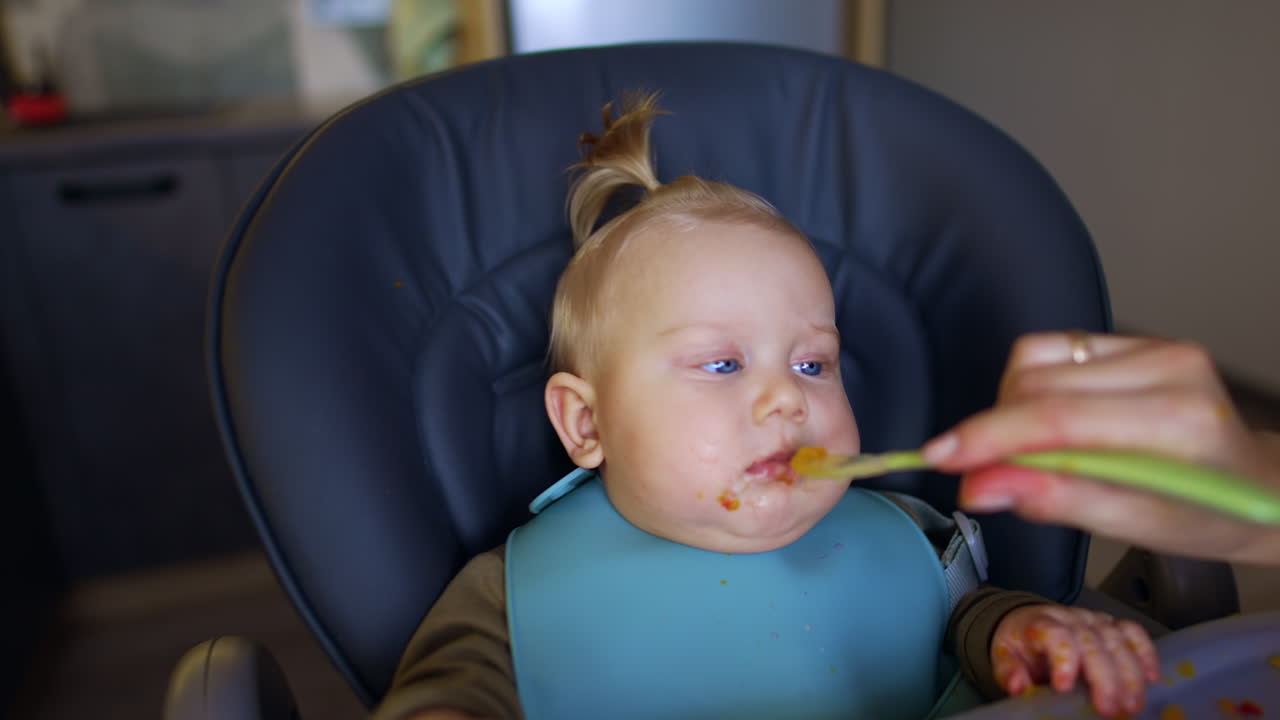 Mommy talks to her adorable baby sitting at high chair. Cute infant child is fed with fresh fruit. Close up.