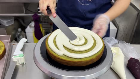 close up commercial kitchen bakery setting shot of a skilful pastry chef using spatula smoothing the buttercream meringue frosting while gently spinning the revolving cake turntable stand