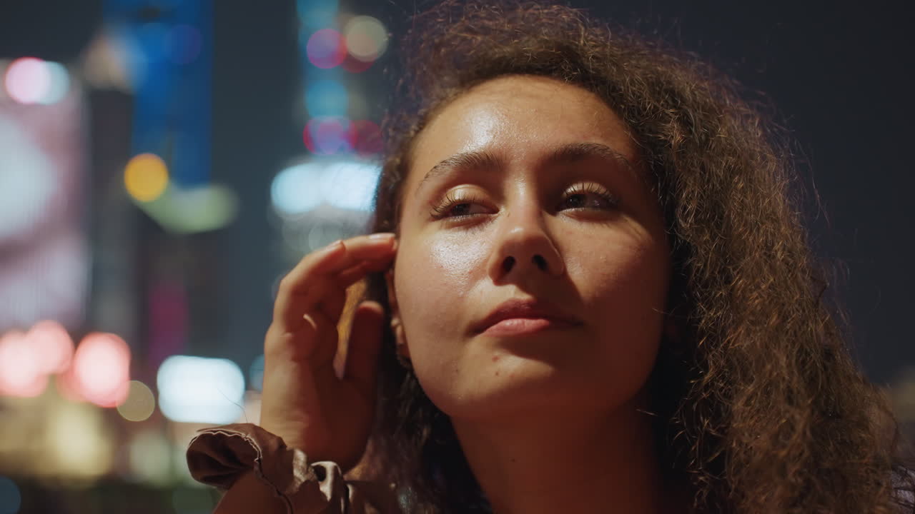 Night city portrait of woman listening to music with neon skyline bokeh, curly hair, wrist bracelets, reflective gaze and ambient glow, rooftop dreamer mood with cinematic slowmotion atmosphere