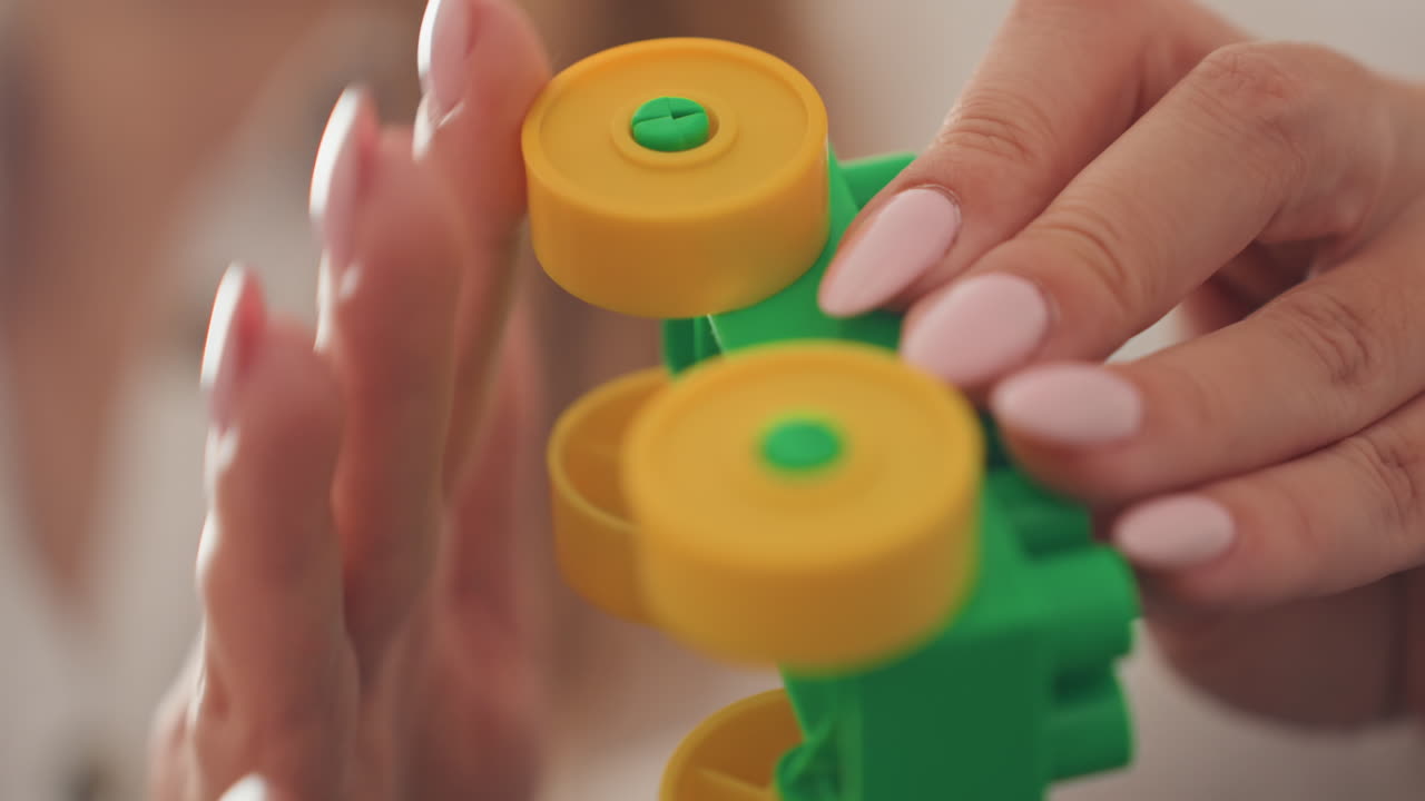 Primo piano mani che giocano con un giocattolo verde giallo, stilista di bellezza donna non specificata che testa la durabilità della manicure ruotando un piccolo giocattolo con ingranaggi di plastica, illuminazione calda e morbida, profondità di campo ridotta, unghia delicata