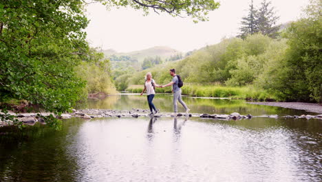 slow motion shot of young couple using stepping stones to cross river whilst hiking in uk lake district