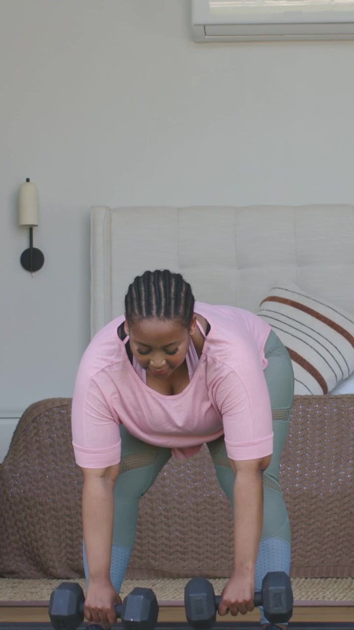 Vertical video: Starting woman in workout top doing hinge row to tone in bedroom with hex dumbbells