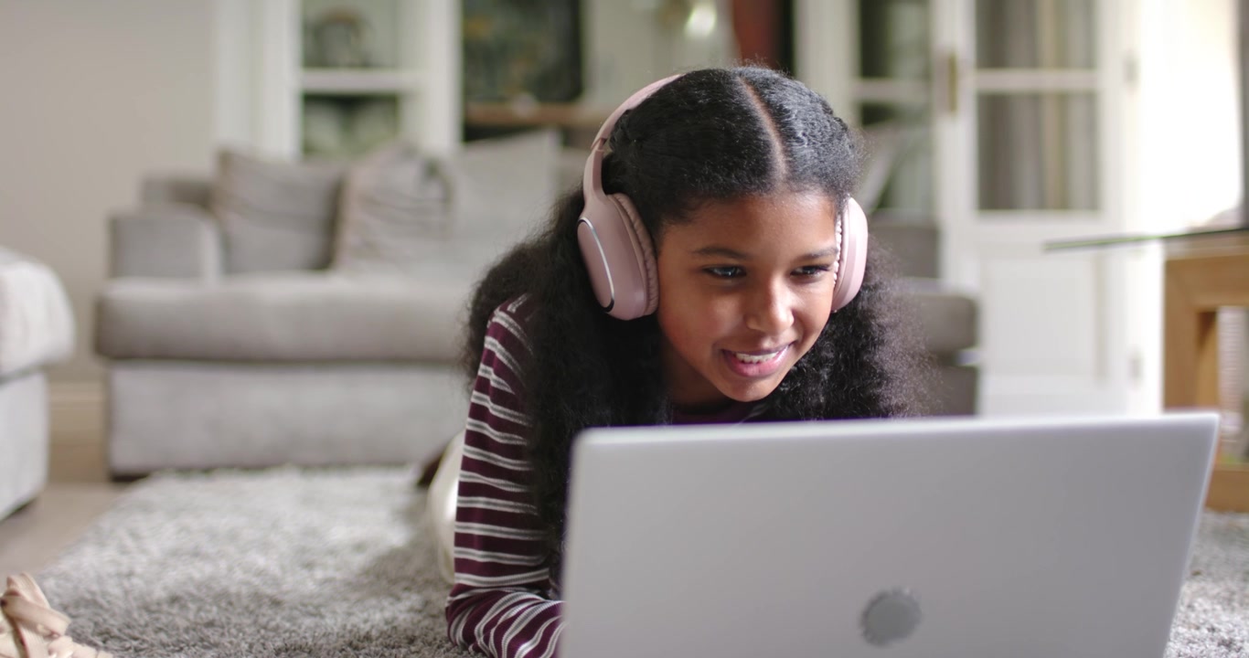 Criança afro-americana inclinando-se e ouvindo uma aula no laptop começando em casa com fone de ouvido rosa