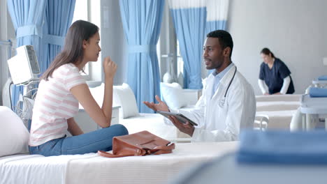 diverse male doctor using tablet and female patient, talking in hospital ward, slow motion