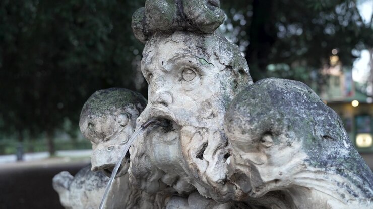 Una scultura di fontana romana a tre teste, macchiata di muschio, lascia scorrere un sottile getto d'acqua dalla bocca centrale nella luce soffusa del giorno
