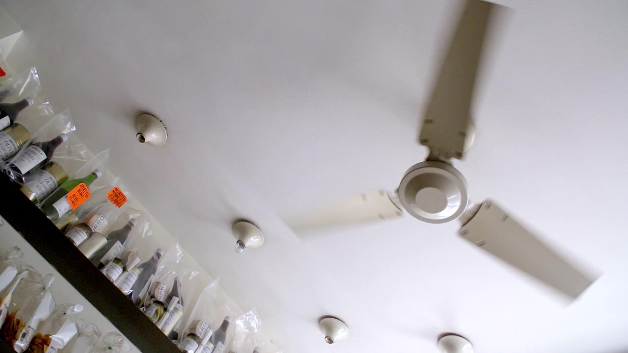 Questa immagine mostra un ventilatore a soffitto in movimento su un soffitto bianco, con diverse luci e scaffali pieni di varie bottiglie, alcune etichettate con adesivi arancioni, in una stanza.