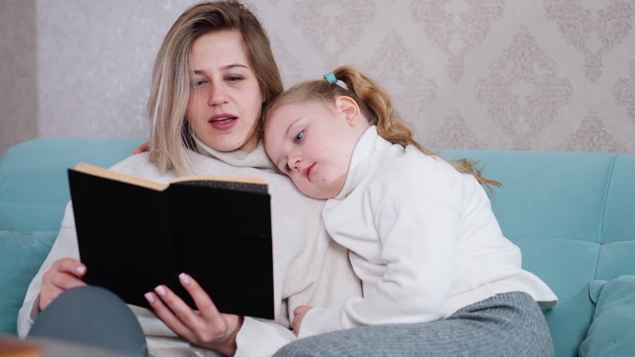 Mother sitting on sofa reading black book aloud while daughter rests head on her shoulder listening attentively, tender family bonding moment full of warmth, comfort, trust, affection, care, parenting