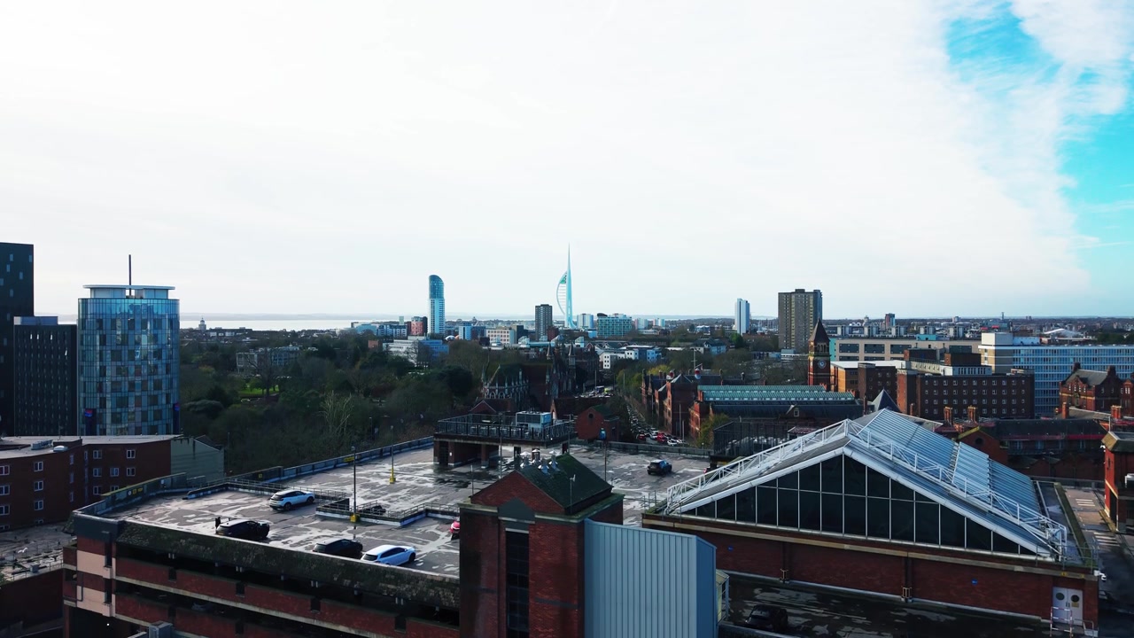 Riprese aeree con drone di un parcheggio multipiano, edifici in mattoni e vetro e una struttura con tetto in vetro a Portsmouth, con una torre in lontananza.