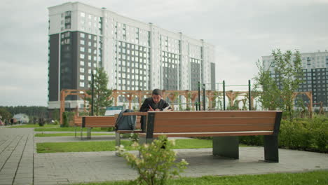 little boy sits bent over wooden bench outdoors studying and writing with concentration, backpack beside him, modern residential buildings in background under cloudy sky