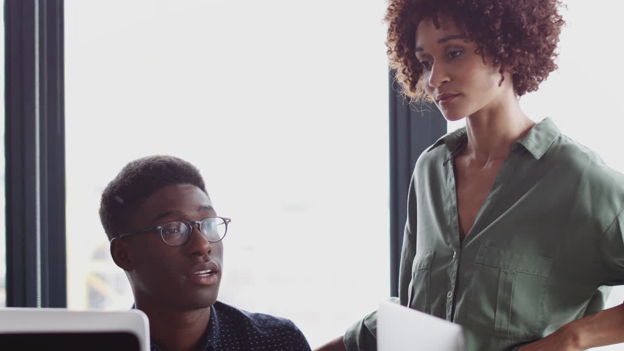 Millennial black woman stands talking to her male creative colleague at his desk, close up