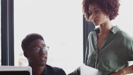 millennial black woman stands talking to her male creative colleague at his desk, close up