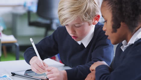 Schoolboy-using-a-tablet-and-stylus-with-a-girl-at-a-desk-in-a-primary-school-class,-close-up