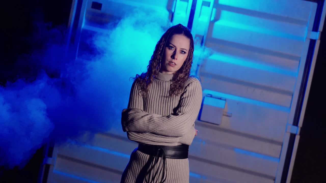 Scatto cinematografico a figura intera di una donna che, di notte, si erge sicura tra fumo blu e nebbia in un contesto urbano industriale. Concept per atmosfere misteriose, immagini di moda e scene di vita notturna suggestive.
