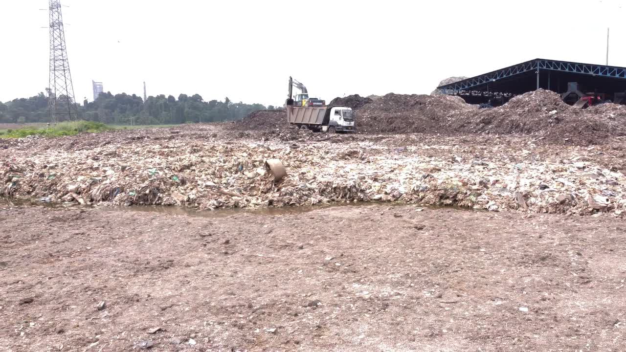 Heavy machinery, excavator, and dump trucks working at a large industrial landfill and waste sorting processing facility, focusing on refuse management and environmental impact