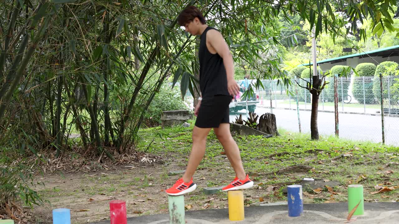 An Asian man in black sportswear balances on colorful stepping stones in a lush park